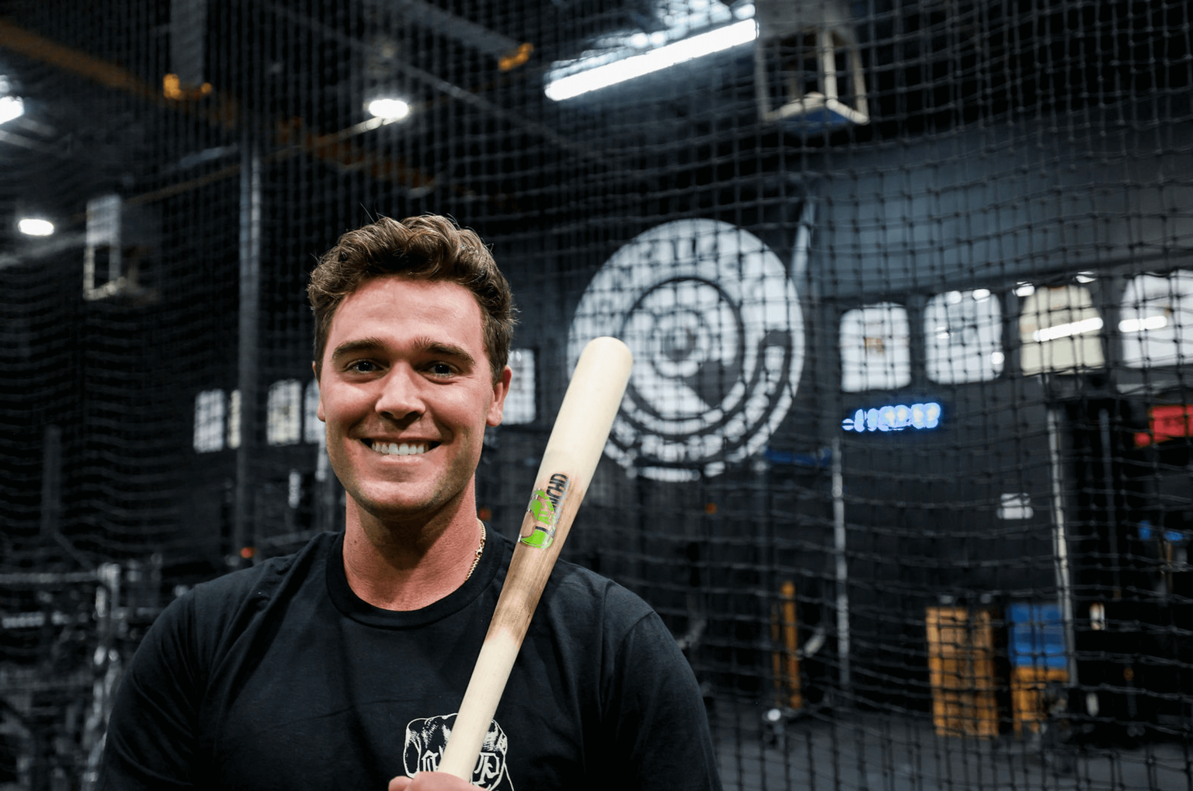 Man holding a baseball bat in an indoor sports facility with a large circular logo on the wall.