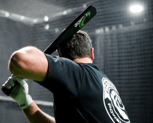 Person in a batting cage holding a baseball bat and wearing a black shirt with a logo.